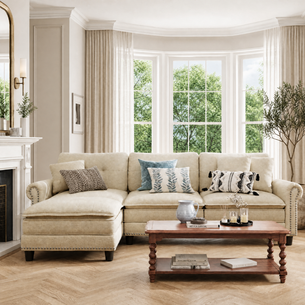 A cream-colored upholstered sectional sofa with nailhead trim, styled in a bright, classic living room with a wooden coffee table, decorative pillows, and large bay windows.